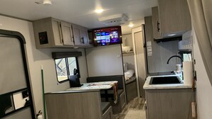 A compact RV interior with a kitchenette, a small dining area, a window, and a TV displaying streaming services.