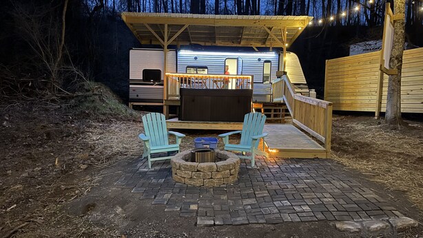 A covered outdoor seating area with two Adirondack chairs, a fire pit, and a wooden deck leading to a camper.