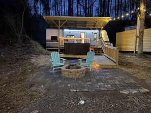 A covered outdoor seating area with two Adirondack chairs, a fire pit, and a wooden deck leading to a camper. - Luxe Mountain Camping with Theater, Hot Tub, Fire Pit & Hiking Trails (Jonesville)