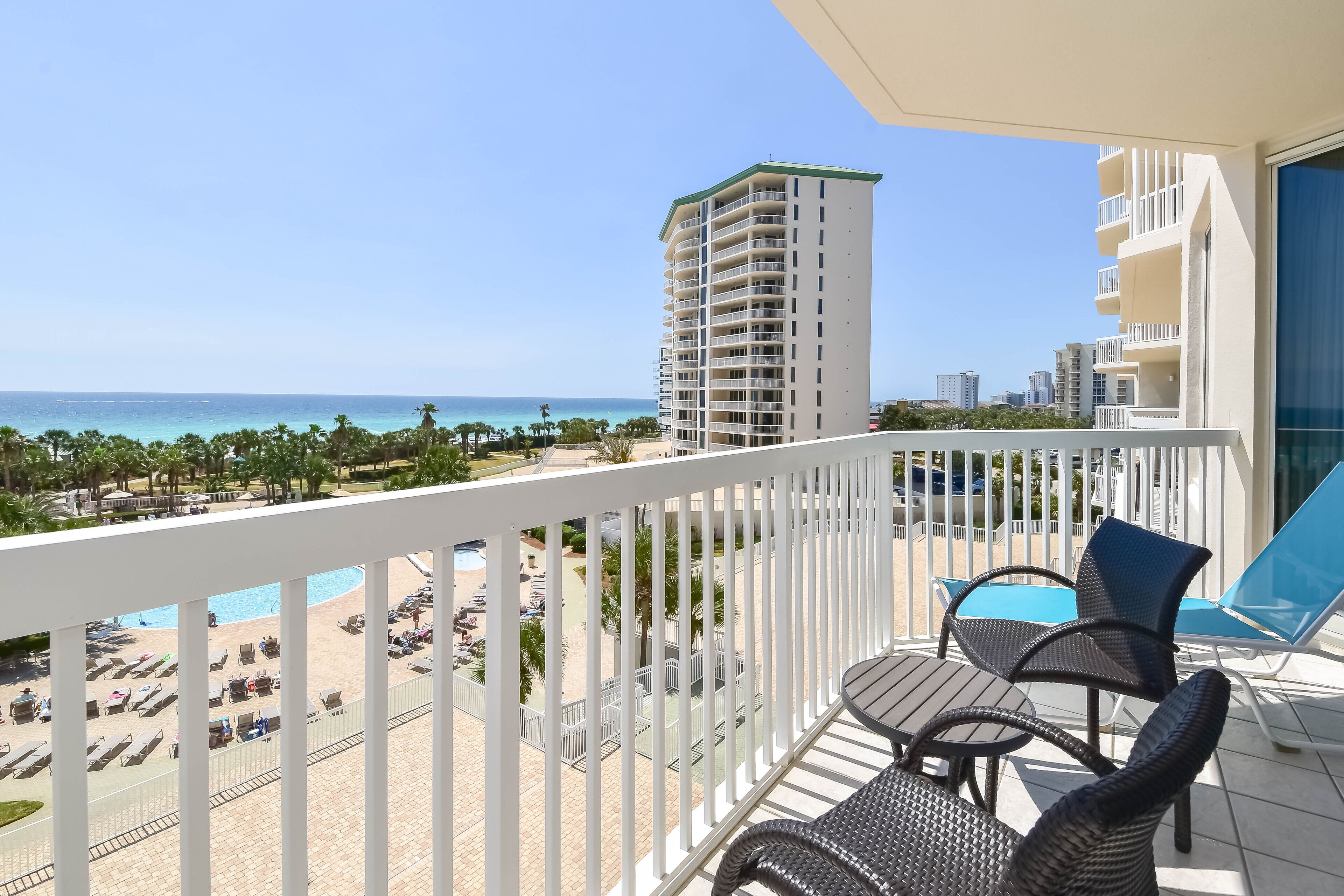 A balcony with wicker chairs and a table, overlooking a pool and beach area with palm trees and a tall building in the background.