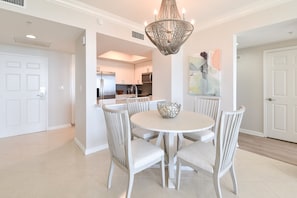 A dining area with a round table and six chairs, a chandelier, and a kitchen in the background. - 5th Floor 2BR Condo at Destin's Silver Shells! (Destin)