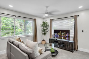 A modern living room with a sofa, a glass coffee table, a TV, and a potted plant. - The Jasmine Apartments by Lowkl (Fort Lauderdale)