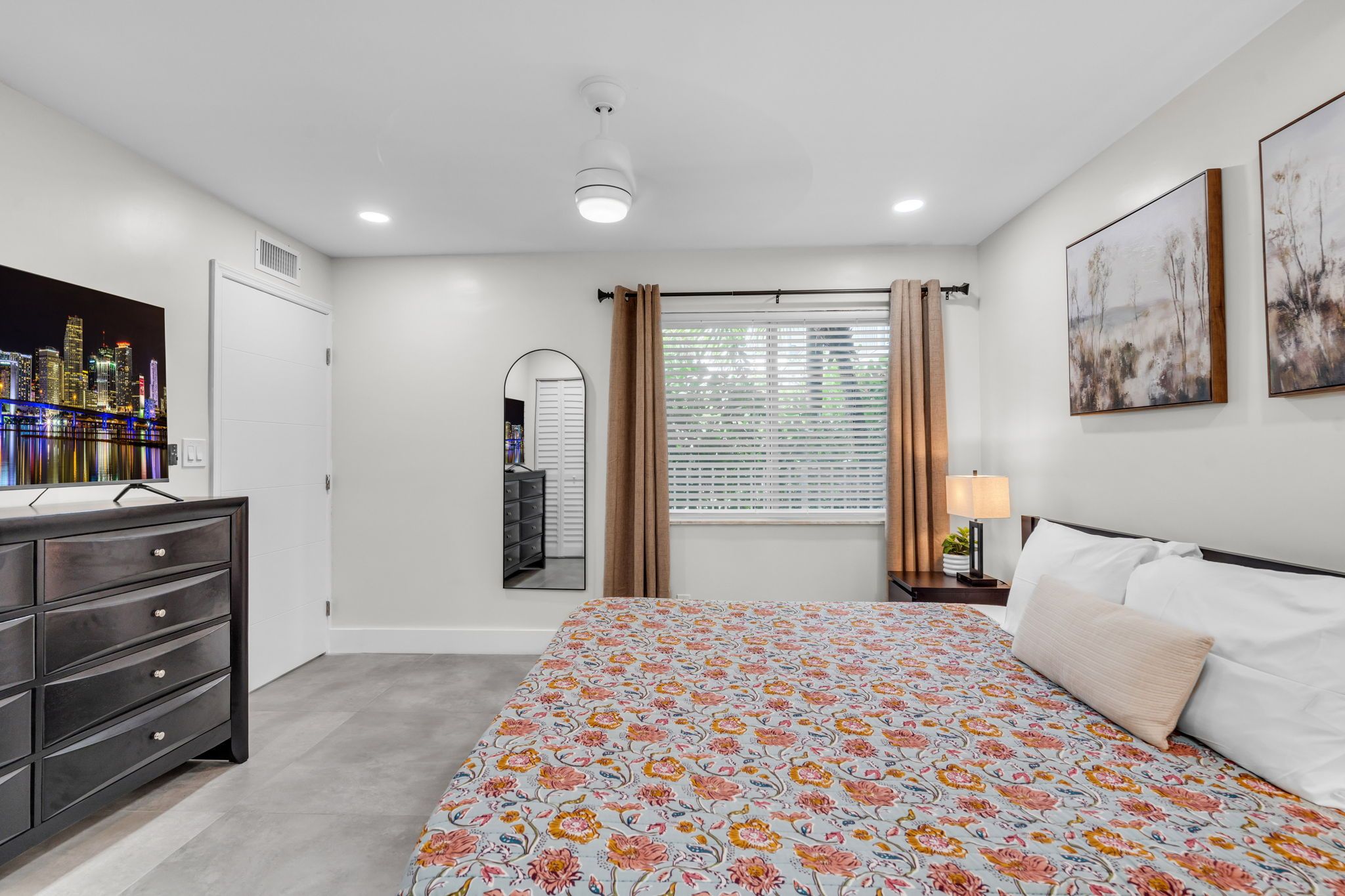 A bedroom with a bed, dresser, mirror, window with curtains, and artwork on the wall.