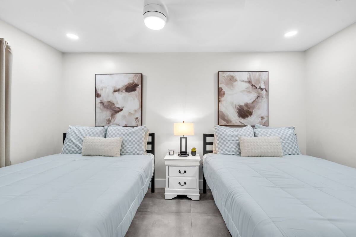 Un dormitorio con dos camas, una mesita de noche con cajones, una lámpara y una obra de arte abstracto en la pared.