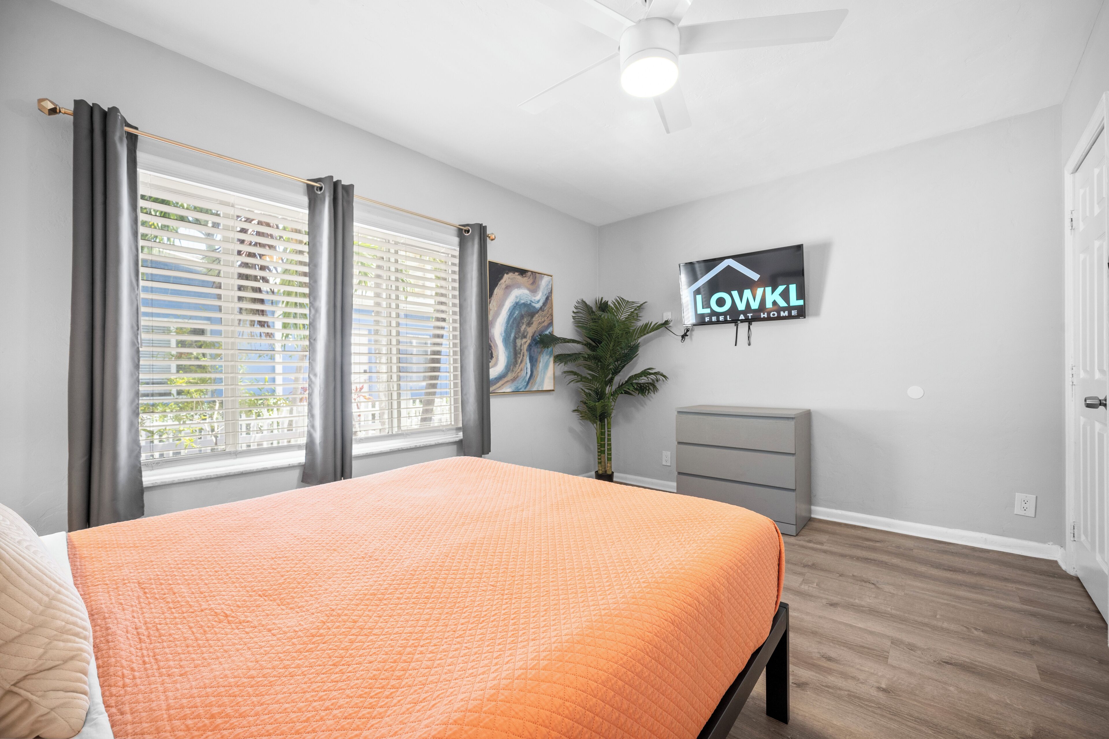 Une chambre à coucher avec un lit, un ventilateur de plafond, une télévision, une plante et une fenêtre avec des stores.