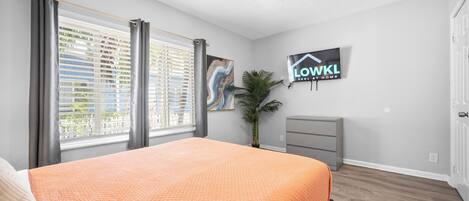 Une chambre à coucher avec un lit, un ventilateur de plafond, une télévision, une plante et une fenêtre avec des stores.