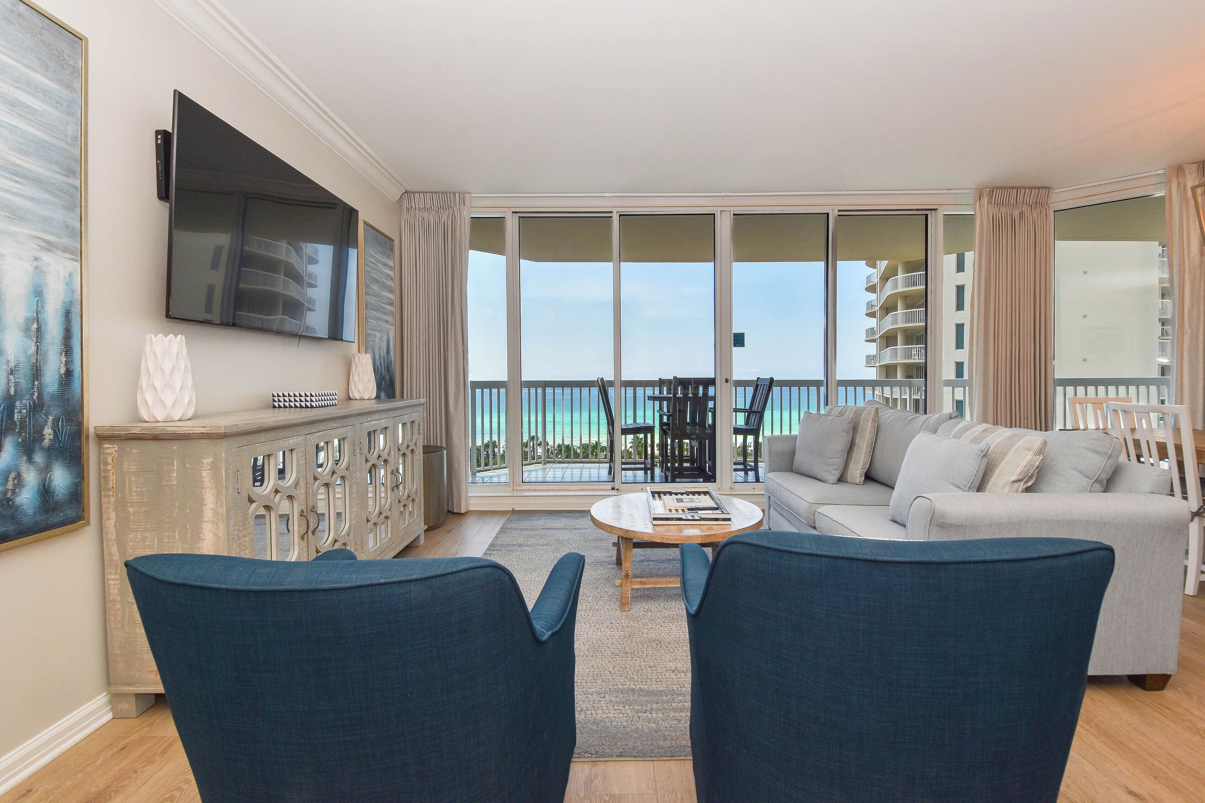 A spacious living room with a large TV, a sofa, and a balcony with a view of the sea.
