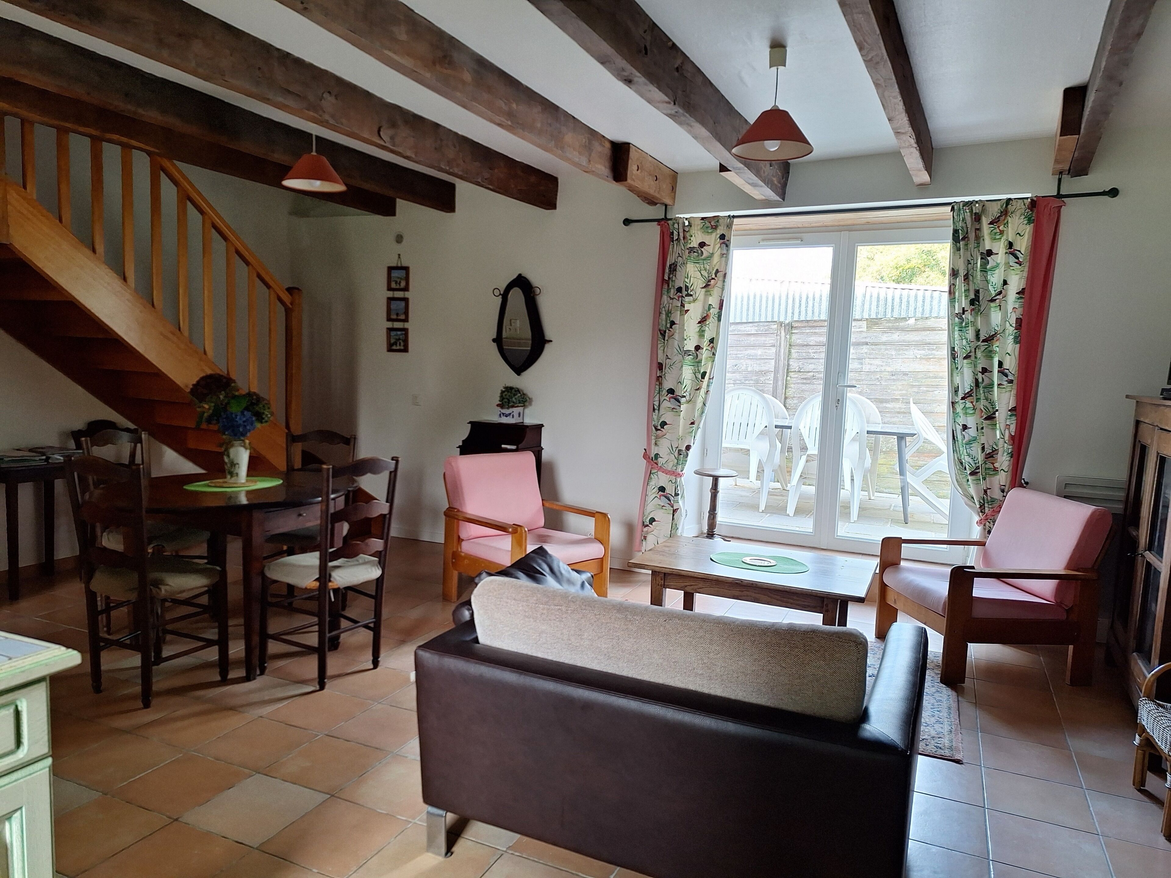 A cozy living room with a wooden staircase, a dining area with a table and chairs, and a large window with curtains.