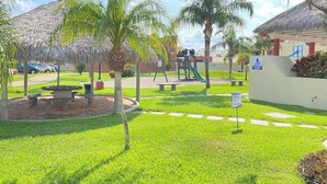 Un espacio exterior cuidado con una zona de estar circular, palmeras, un parque infantil y un edificio con techo de paja.