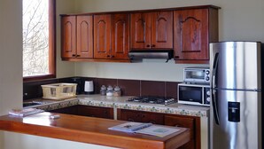 A kitchen with wooden cabinets, a stainless steel refrigerator, a microwave, a toaster, and a window with a view of trees. - Galeodan Dorado Suite - Spacious Family Suite with Gorgeous Ocean View (Puerto Baquerizo Moreno)