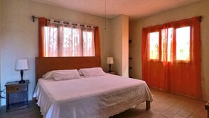 A bedroom with a bed, a nightstand, a lamp, and two windows with curtains. - Galeodan Dorado Suite - Spacious Family Suite with Gorgeous Ocean View (Puerto Baquerizo Moreno)