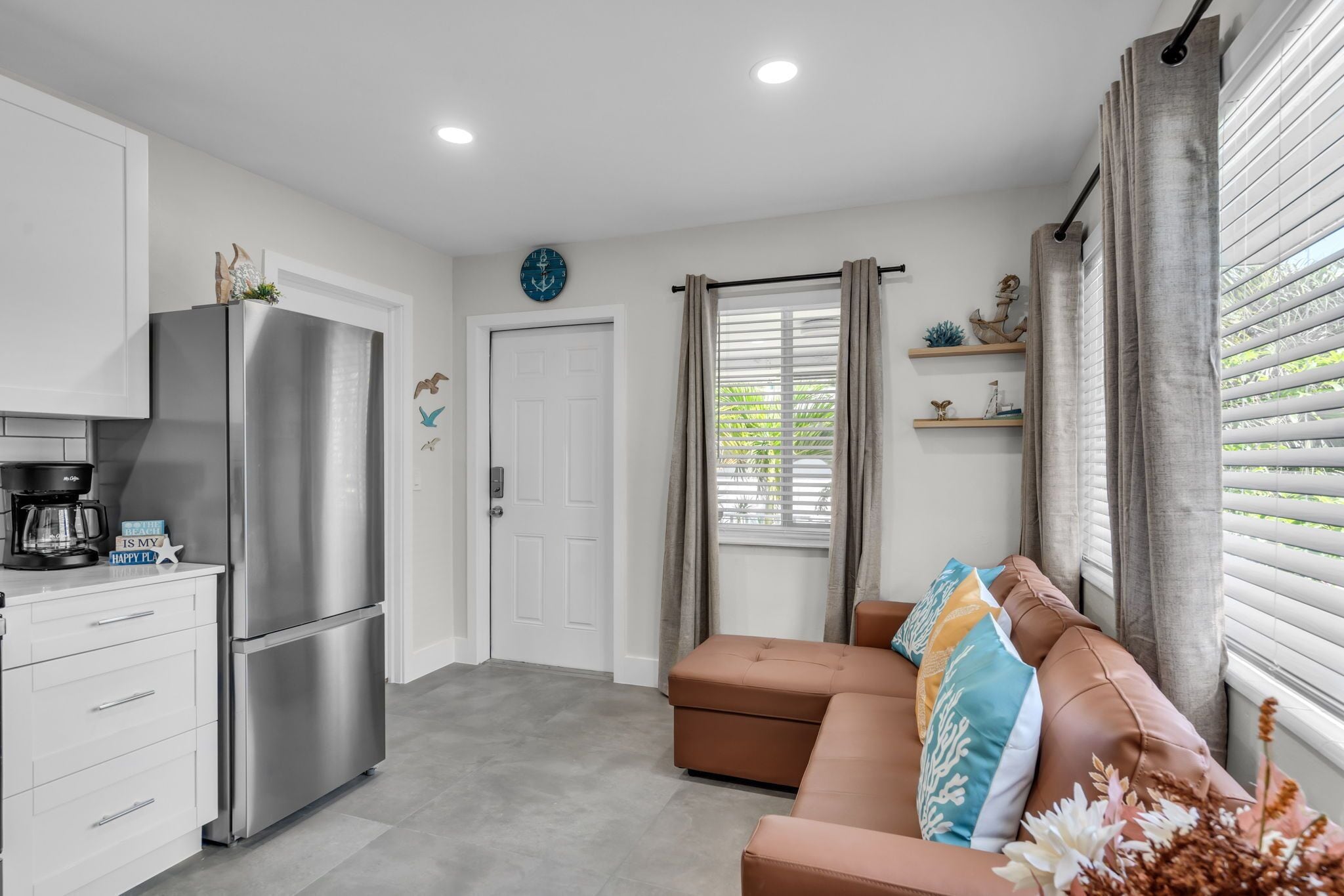 A compact living space with a kitchenette, a sofa with decorative pillows, a coffee maker, and a view through a window with blinds.