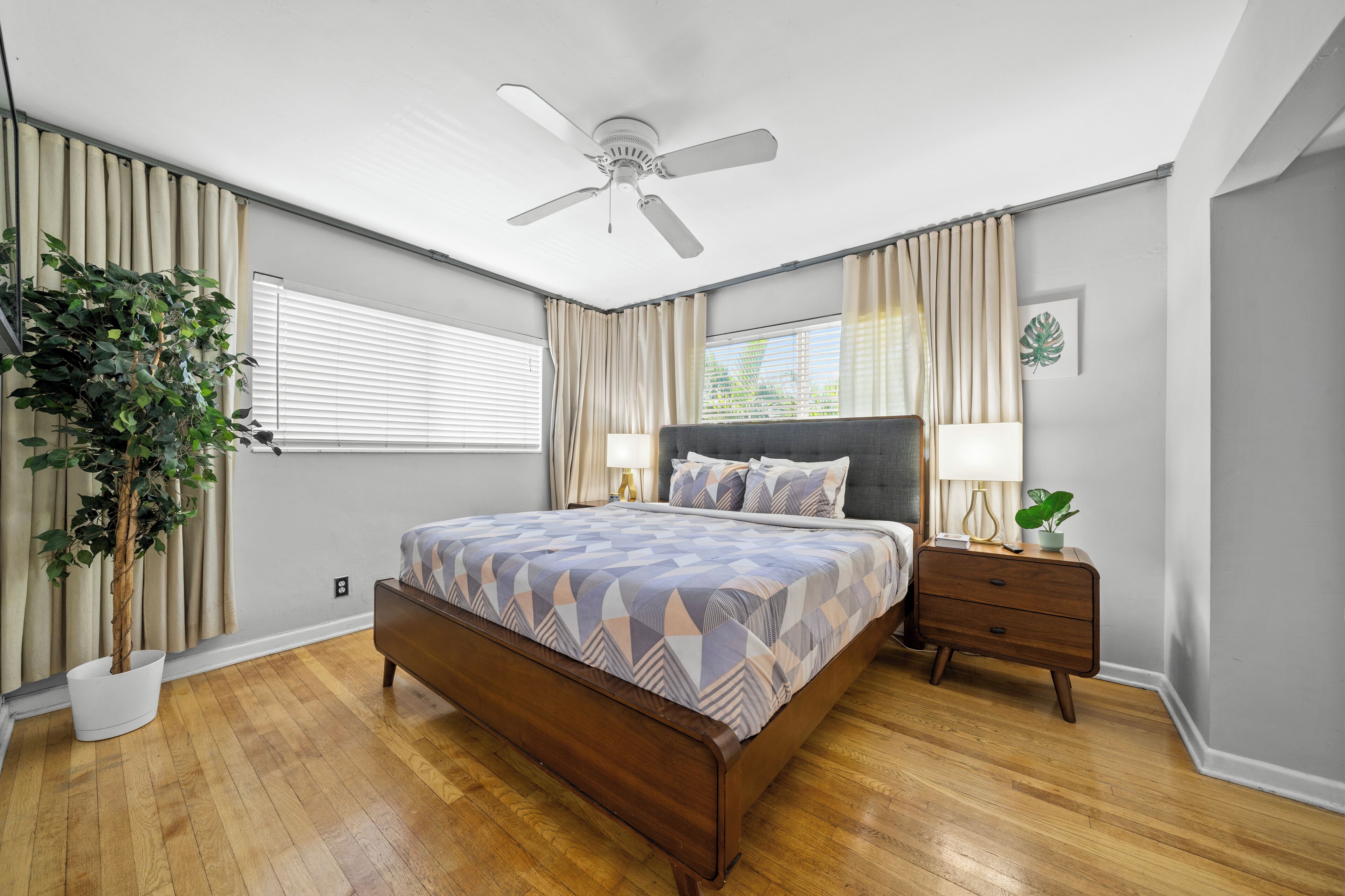 A bedroom with a wooden bed, a ceiling fan, a bedside table with a lamp, and a potted plant.
