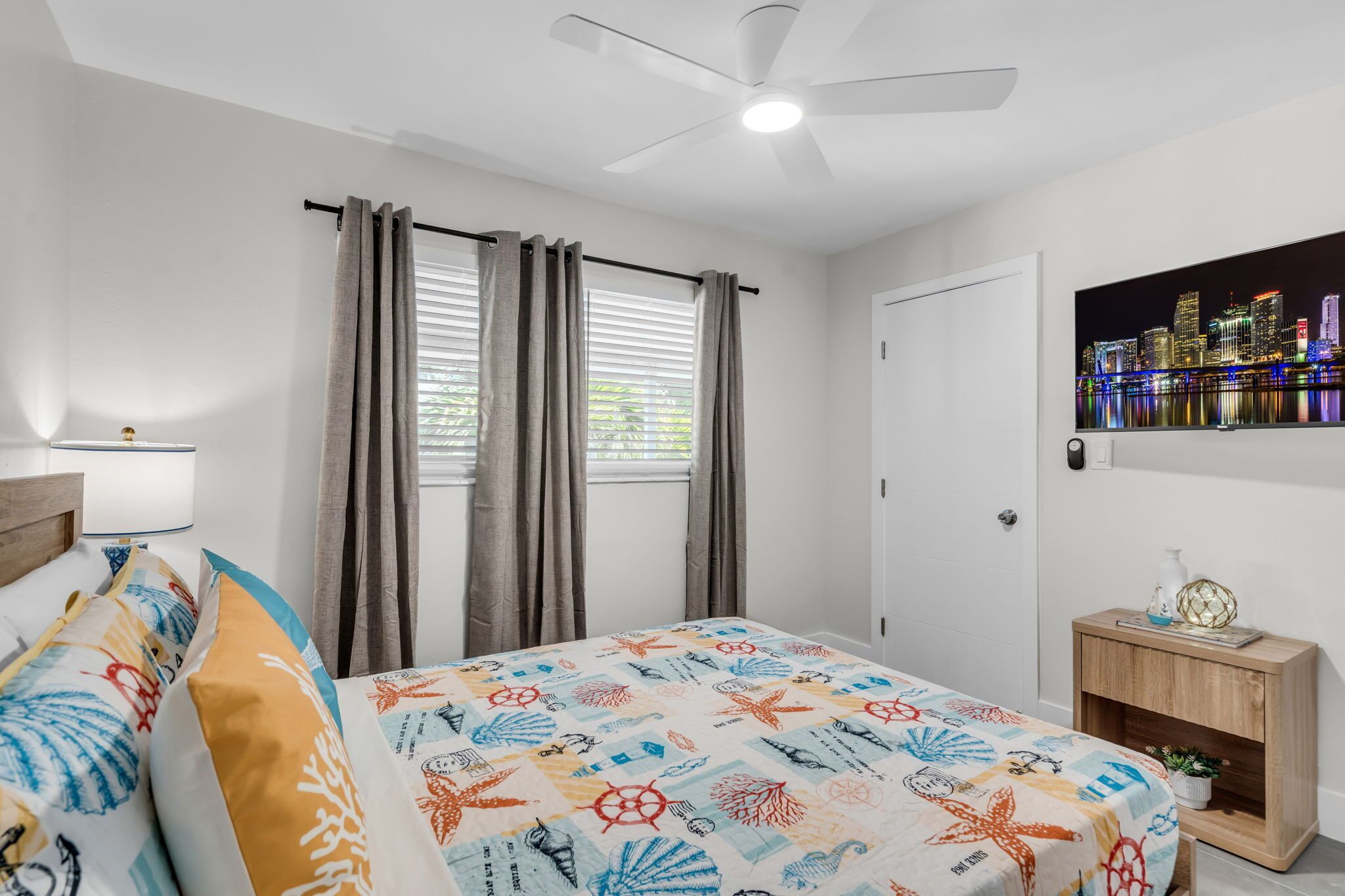 A bedroom with a bed, curtains, a ceiling fan, a nightstand, and a cityscape painting.