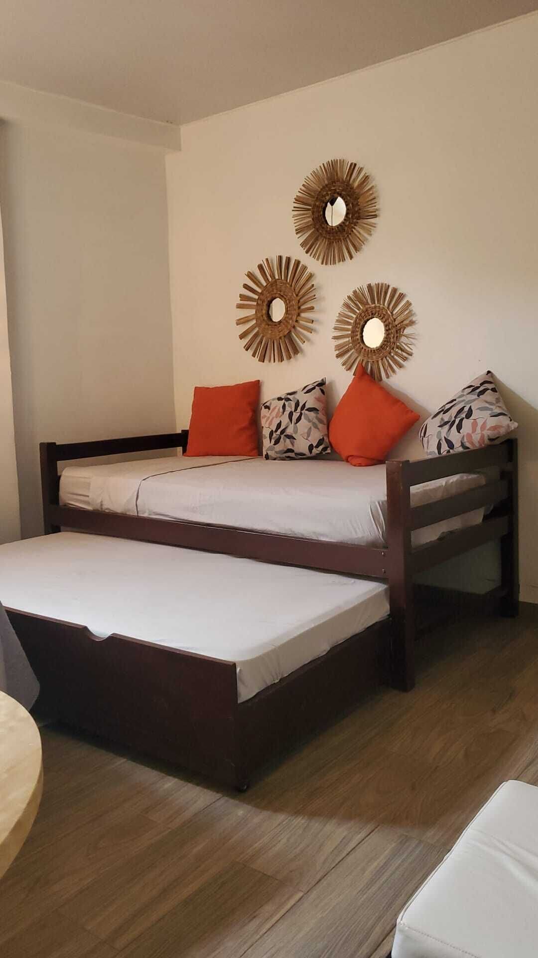 Un lit double avec un cadre en bois, deux oreillers orange et un coussin à motifs. Il y a trois miroirs décoratifs fixés au mur. La pièce est dotée d’un plancher en bois et de murs blancs.