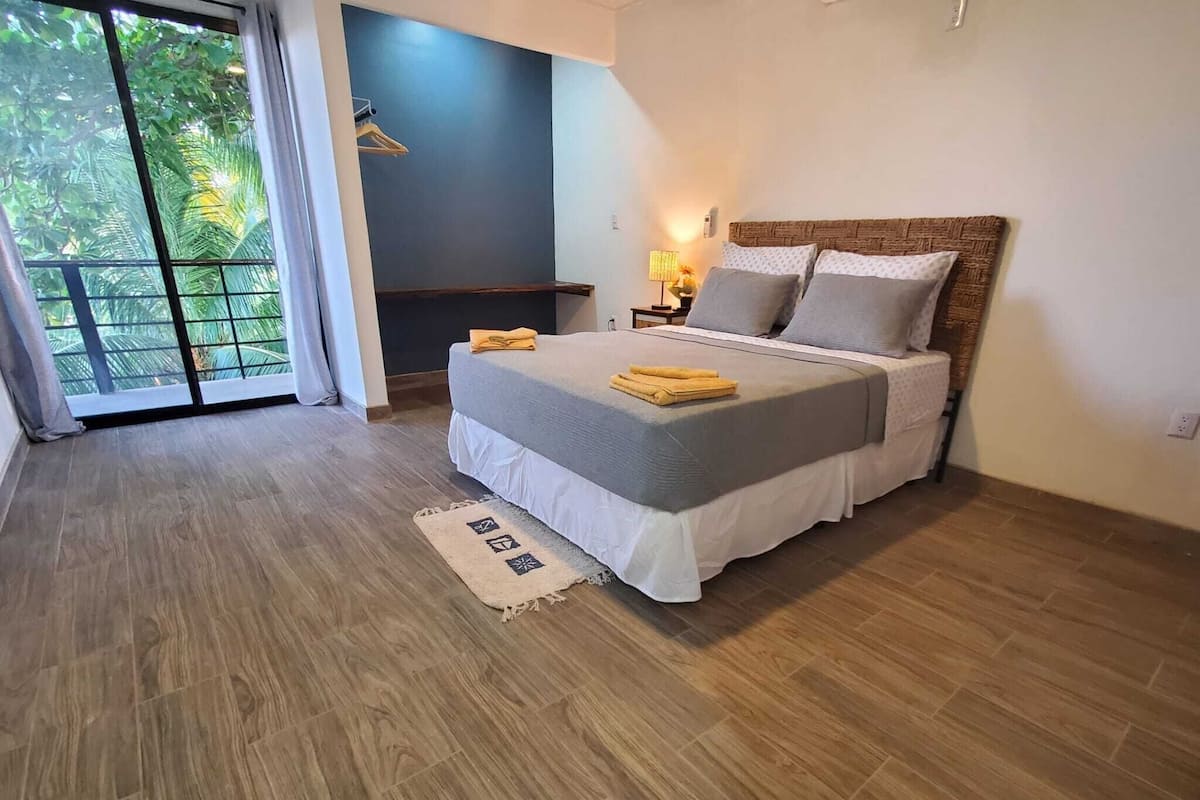 A bedroom with a bed, a nightstand, a lamp, a window with a view of trees, and a door leading to a balcony.