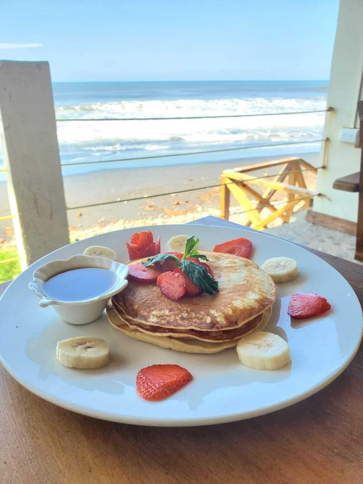 Ein Frühstücksteller mit Pfannkuchen, Erdbeeren, Bananen und Sirup auf einem Tisch mit Blick auf Strand und Meer.