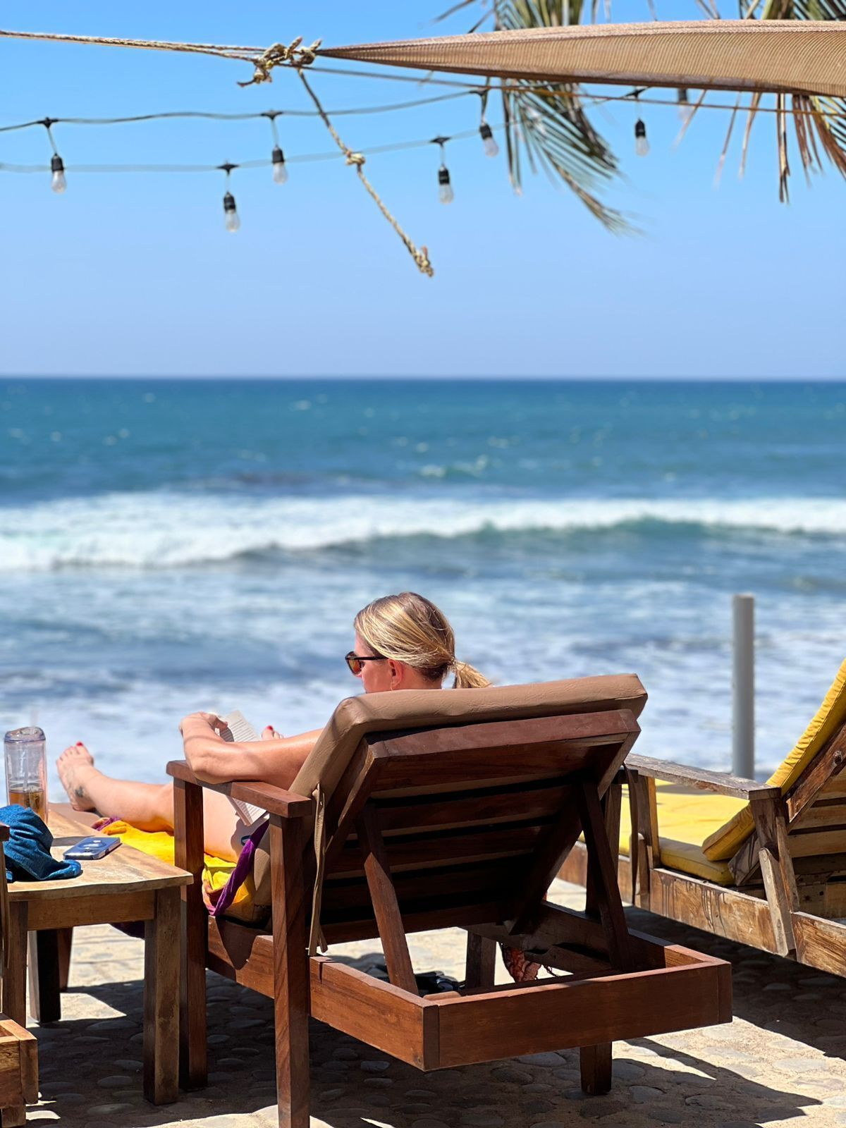Un entorno frente al mar con una silla de madera, una mesa con una bebida y vista al océano.