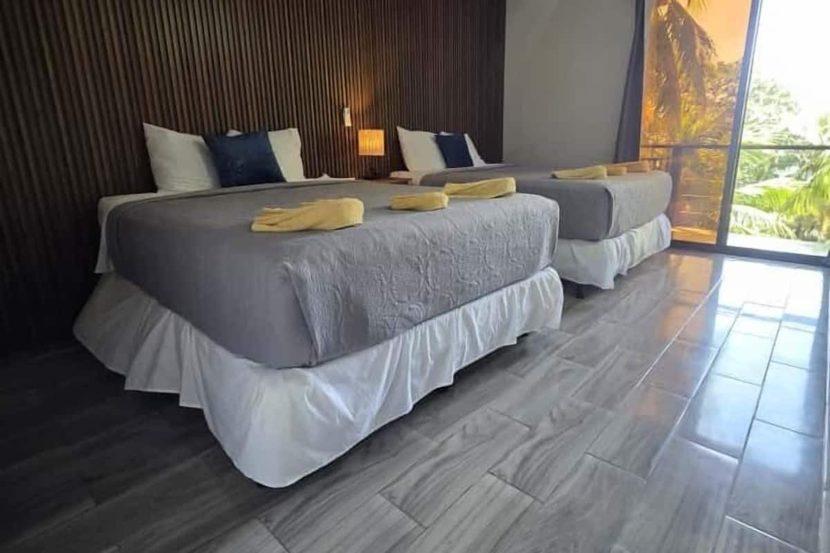 A modern hotel room with two beds, a wooden wall, and a large window with a view of greenery.