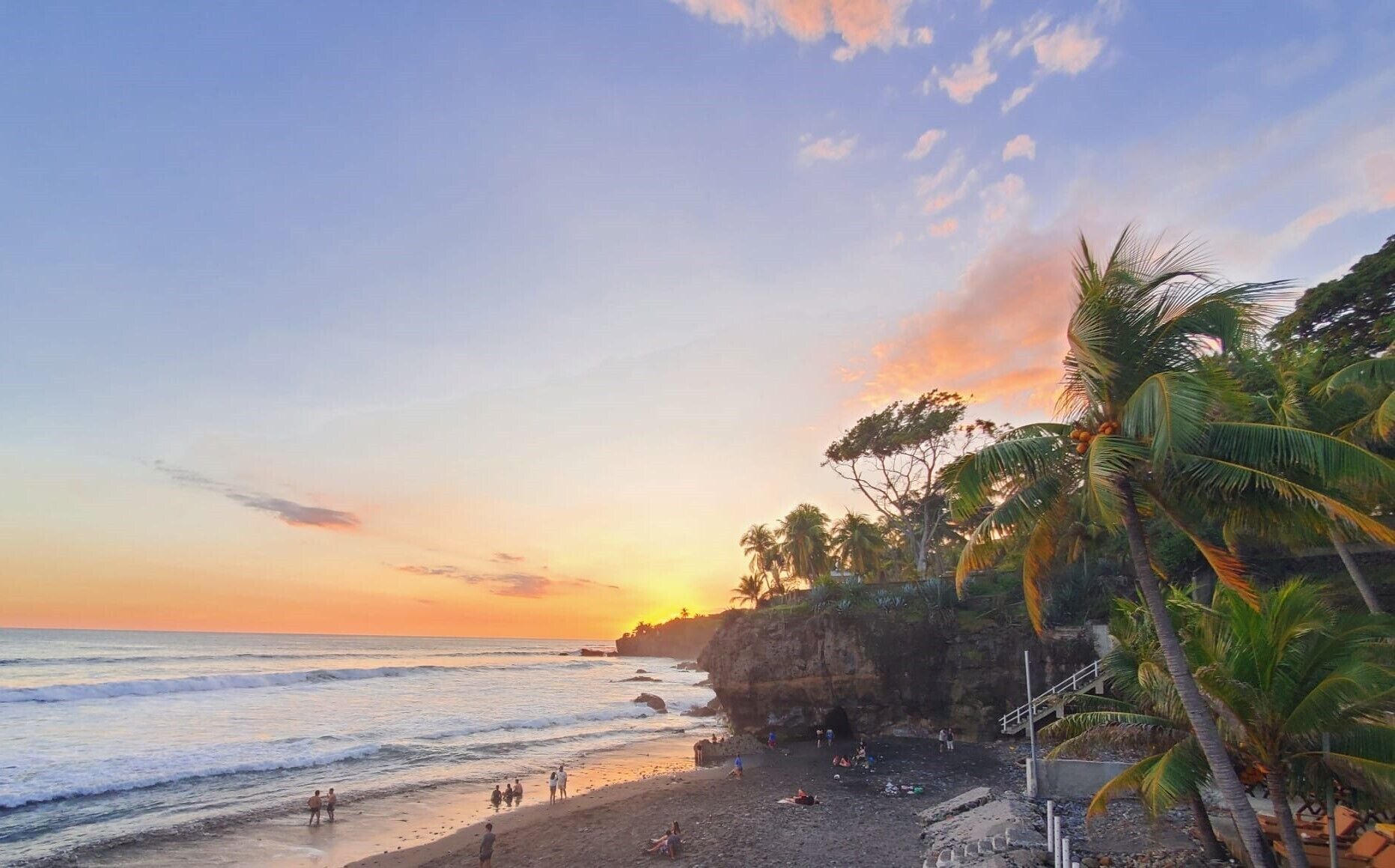 A beach with palm trees, rocky cliffs, and people enjoying the sunset.