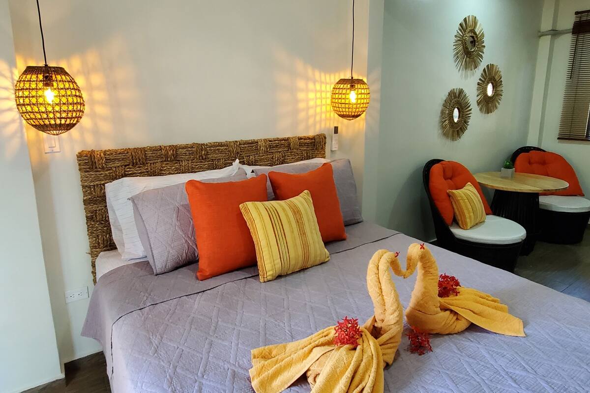 A neatly made bed with a towel swan decoration, a woven headboard, and two hanging lamps.