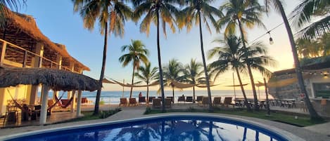 A poolside area with palm trees, lounge chairs, and a thatched-roof structure.