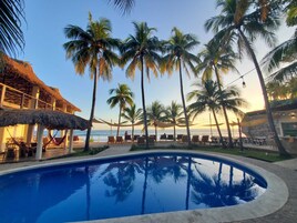A poolside area with palm trees, lounge chairs, and a thatched-roof structure.