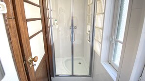 Un box de ducha con paredes de vidrio, suelo de baldosas blancas y ventana a la derecha.