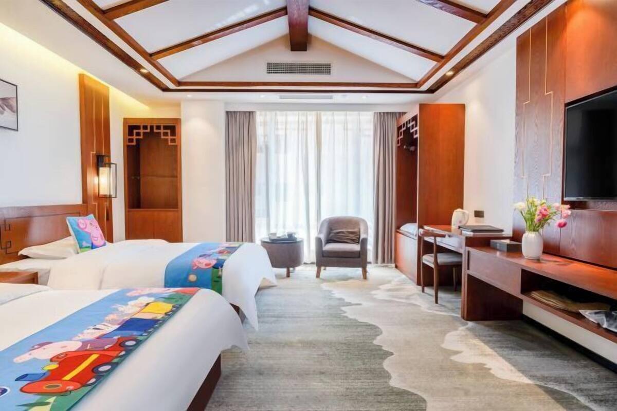 Una habitación de hotel con dos camas, un escritorio, una silla, un televisor y un ventanal con cortinas.