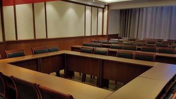A conference room with a long table and multiple chairs, a large window with curtains, and a plain wall with a panel design.