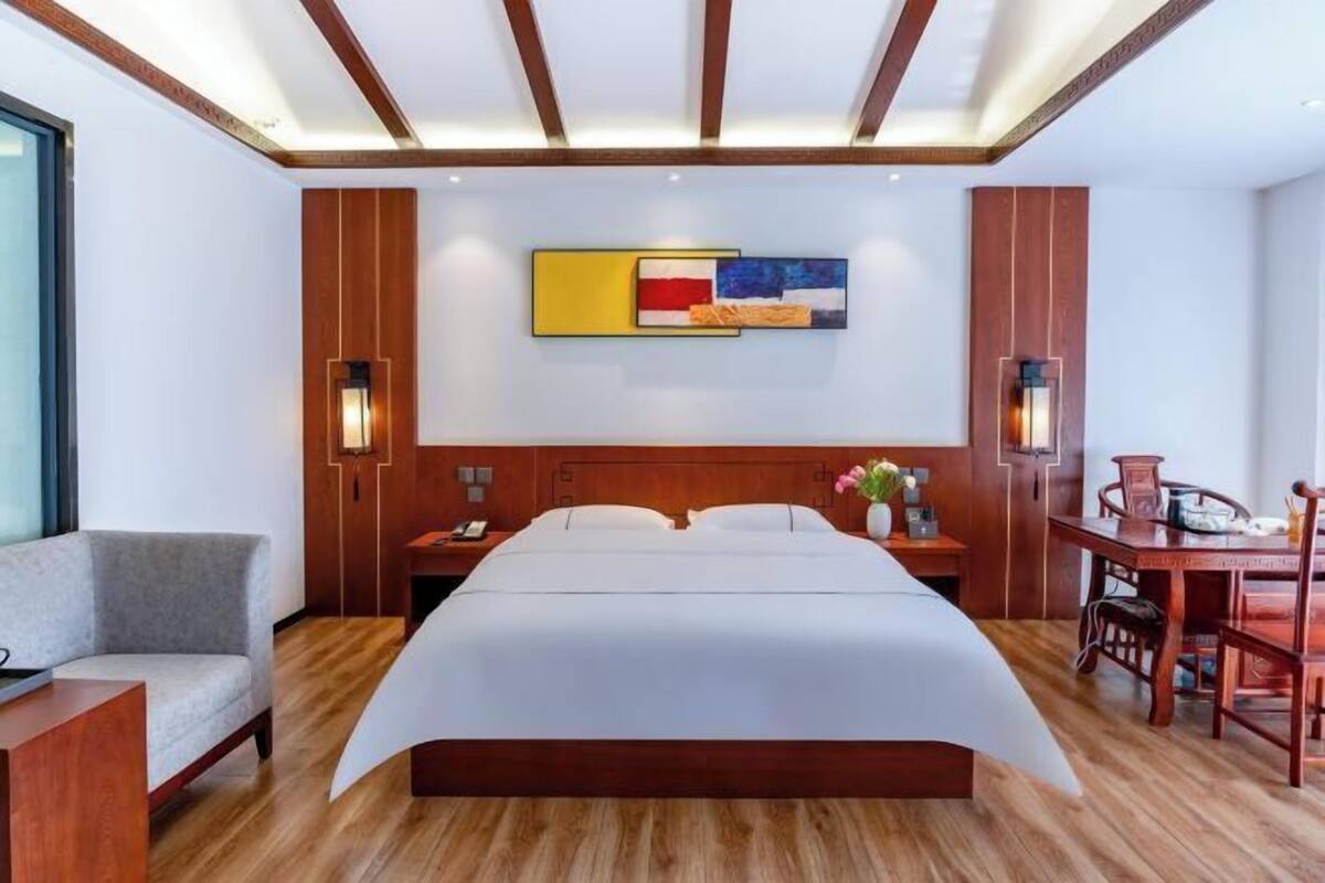 Habitación de hotel con cama de cabecera de madera, un escritorio con silla, un sofá y una obra de arte colgada en la pared.