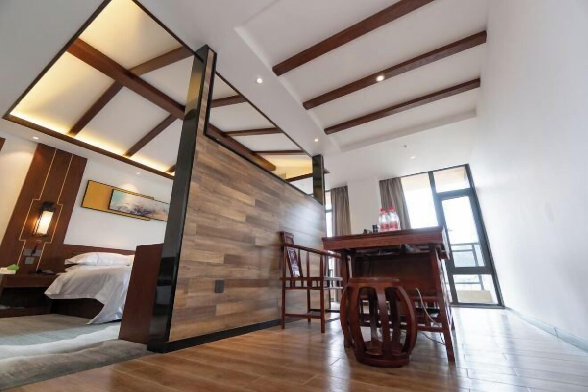 Een moderne hotelkamer met een houten vloer, een bed, een bureau en een stoel. Er is een groot raam dat zorgt voor natuurlijk licht.