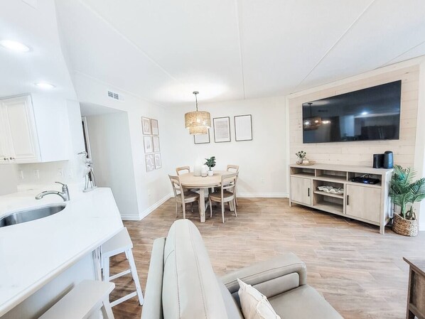 Une cuisine moderne avec des meubles blancs, un coin repas avec une table ronde et des chaises, un grand téléviseur à écran plat et un plancher en bois.