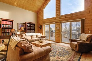 A spacious living room with a high ceiling, wooden walls, and large windows. There are two sofas, a coffee table, and a bookshelf filled with books. - Game Room + Hot Tub! Rocky Mountain Escape (Fairplay)