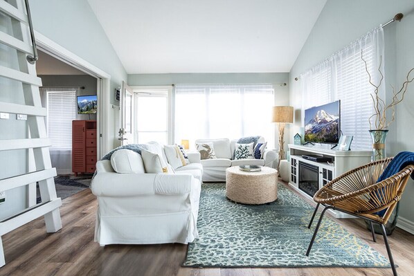 A living room with a white sofa, a round ottoman, a wicker chair, a television, and a fireplace.