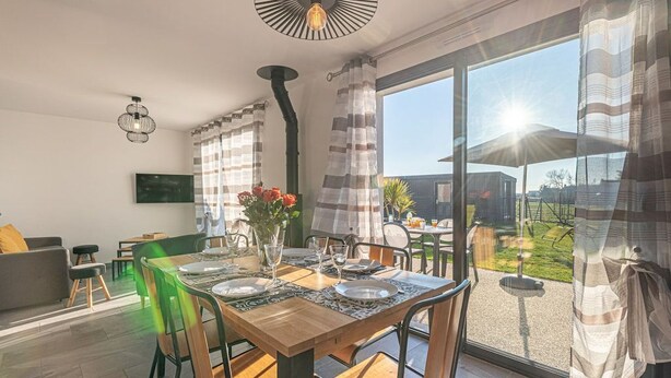 Un coin repas avec une table en bois dressée pour quatre personnes, entourée de chaises. On y trouve une grande fenêtre avec des rideaux, un téléviseur fixé au mur et un luminaire au plafond.