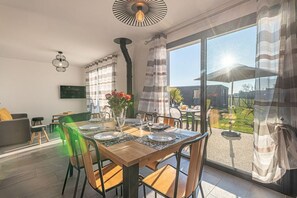 Un coin repas avec une table en bois dressée pour quatre personnes, entourée de chaises. On y trouve une grande fenêtre avec des rideaux, un téléviseur fixé au mur et un luminaire au plafond.