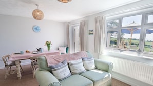 A cozy room with a green sofa, a dining table with chairs, a clock, and a view of the outdoors through large windows. - The Seaside Inn (Seaton)