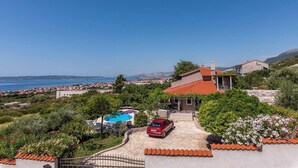 Une propriété située sur la côte, dotée d’une piscine, avec une voiture rouge garée devant, et offrant une vue sur la mer et une ville lointaine.