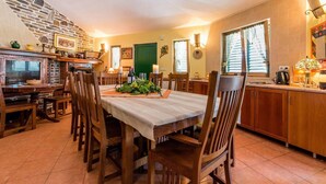 Un coin repas avec un mur en pierre, des chaises en bois, une table recouverte d’une nappe blanche et un espace cuisine doté de meubles en bois et d’une fenêtre.