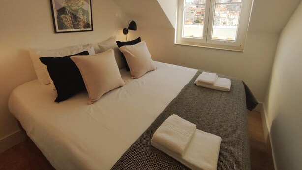 A bedroom with a bed, pillows, a blanket, a framed picture, a lamp, and a window with a view of buildings. - Lisbon Skyline Apartment by Misha s Place 5E (Lisbon)