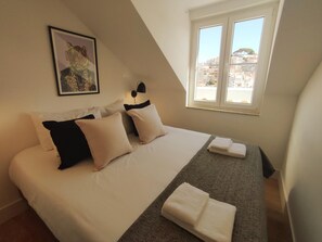 Quarto com cama, almofadas, cobertor, quadro emoldurado, abajur e janela com vista para edifícios.