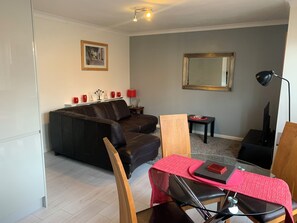 Ein Wohnzimmer mit einem dunkelbraunen Ledersofa, einem Holzstuhl, einem Glastisch mit roter Tischdecke und einem kleinen Beistelltisch mit Lampe und roter Vase.