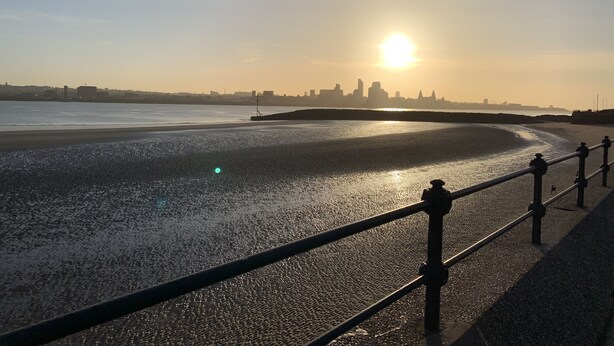 Vista de um pôr do sol à beira-mar, com o horizonte da cidade ao longe, uma praia arenosa e um corrimão de metal na orla.