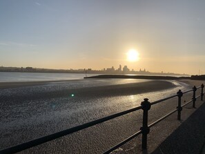 Ein Küsten-Sonnenuntergang mit Stadtansicht in der Ferne, einem Sandstrand und einer Metallgeländer entlang der Küste.