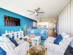 Ein modernes Wohnzimmer mit blauer Akzentwand, Deckenventilator, Flachbildfernseher, blauem Sofa, Korbmöbeln und einer Küche im Hintergrund.