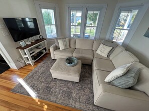 A living room with a sectional sofa, a coffee table, a flat-screen TV, and a window with a view of the outside. - Modern Farmhouse with Porch near Lake & Downtown (Green Lake)
