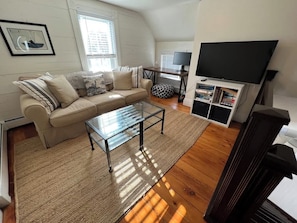 A living room with a sofa, glass coffee table, TV, and bookshelf. - Modern Farmhouse with Porch near Lake & Downtown (Green Lake)
