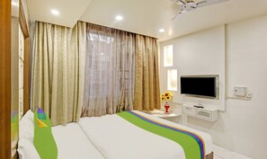 A hotel room with a bed, curtains, a TV, a vase with sunflowers, and a ceiling fan. - Treebo Jannat Residency (New Delhi)