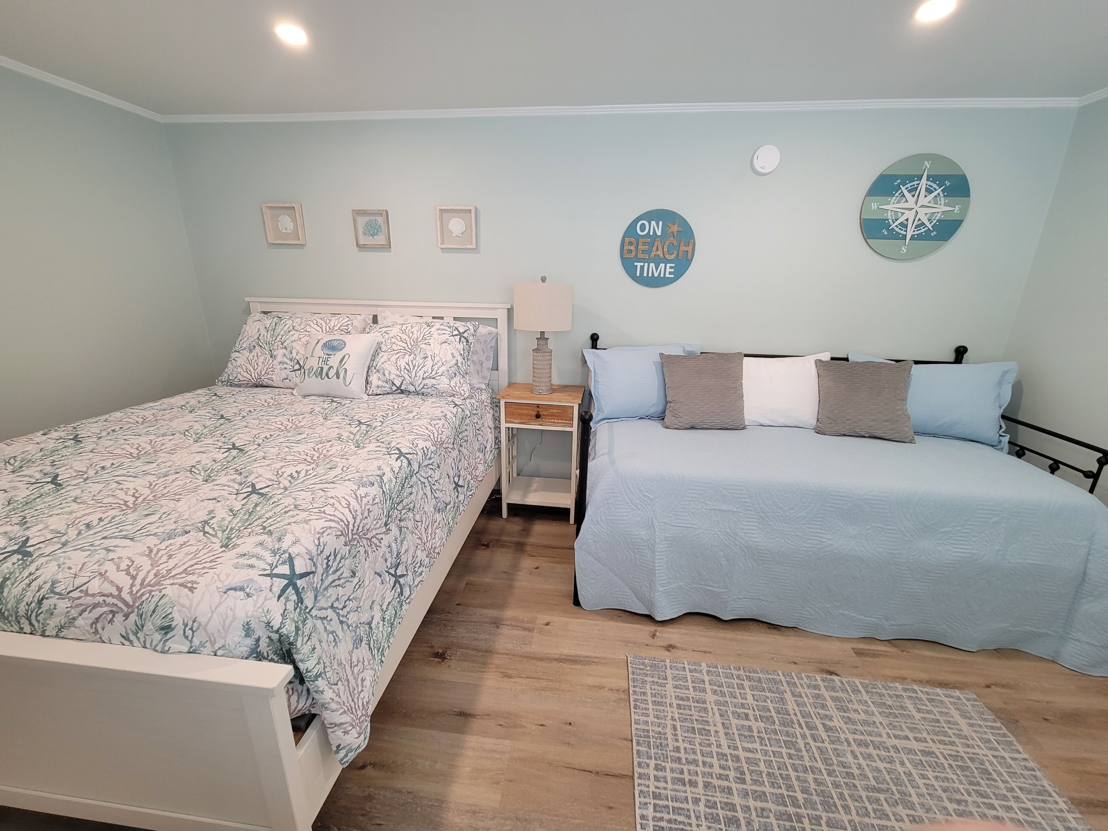 A bedroom with two beds, a nightstand, a lamp, and wall decorations including a compass and a sign saying "ON BEACH TIME".